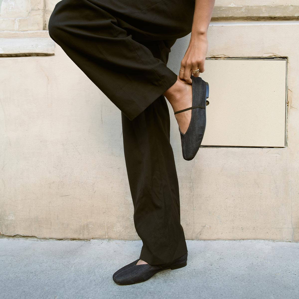 A person in black pants stands against a beige wall, one leg crossed, adjusting The Phoebe - Indigo Denim flat by margaux-development on their foot.