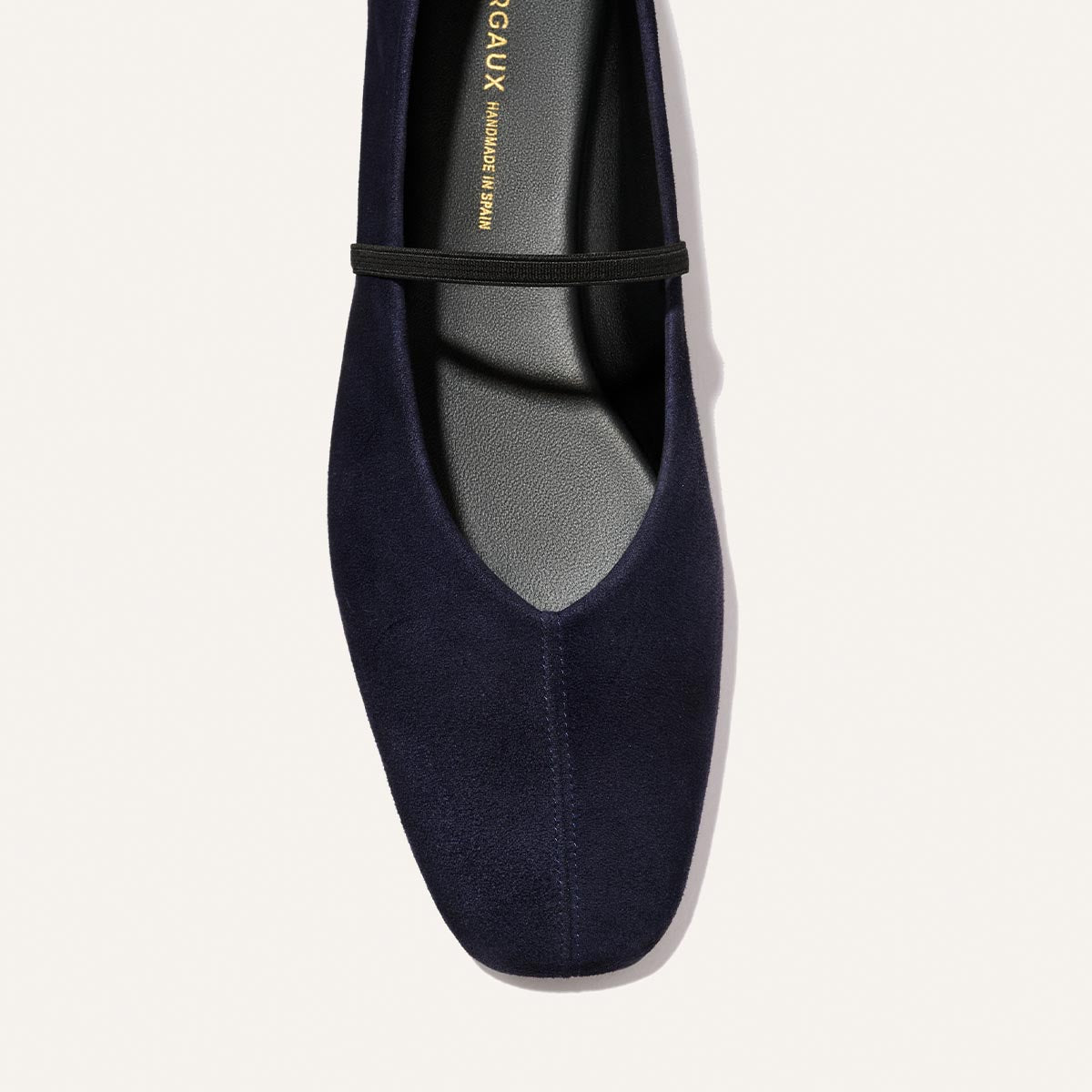 A close-up of Margaux’s The Phoebe in Indigo Suede, a navy blue suede flat featuring a black strap across the top, photographed from above on a light background. Crafted with sacchetto construction, this deconstructed flat combines elegance and exceptional comfort.