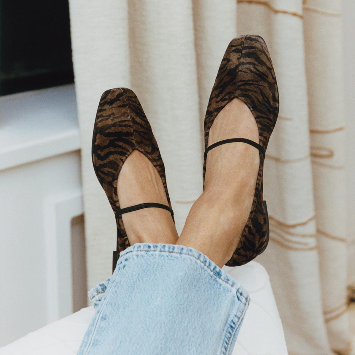 A person wears light blue jeans and Margaux's The Phoebe - Brown Zebra Suede Mary Jane flats, featuring black straps and supple sacchetto construction, as they rest their feet on a white surface with beige curtains behind.