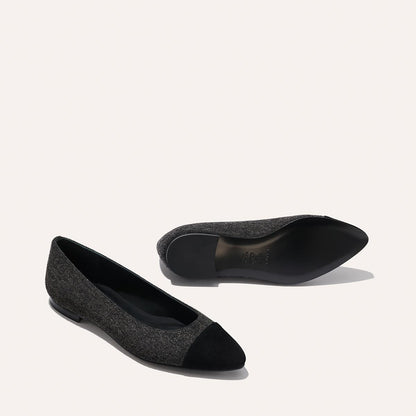 Two The Pointe - Grey Flannel flats by margaux-development are displayed on a light background, featuring dark gray uppers with black pointed toe caps. One flat stands upright, while the other lies on its side to reveal the sole.
