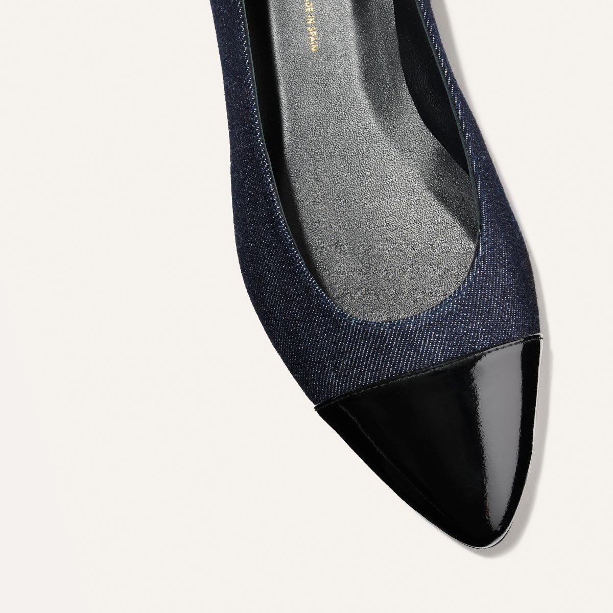 A close-up of The Pointe - Indigo Denim and Patent by margaux-development features a sleek dark blue denim flat with a shiny black patent pointed toe, displayed on a light background.