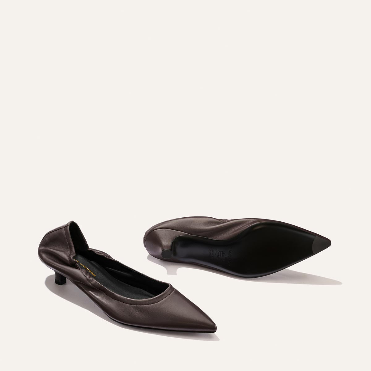 A pair of The Roma 40 - Espresso Nappa kitten heel shoes by Margaux, one standing upright and the other lying on its side, displayed on a light background and embodying timeless elegance with their chic, effortless sophistication.