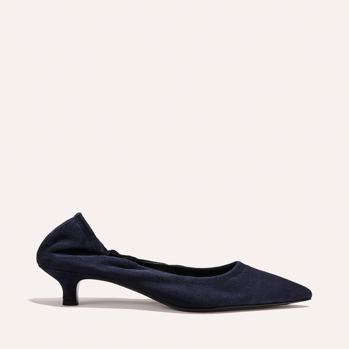 The Roma 40 - Indigo Suede by Margaux is a navy blue suede pump with a low, walkable kitten heel and ruched back detail, showcasing an elegant silhouette against a plain, light background.