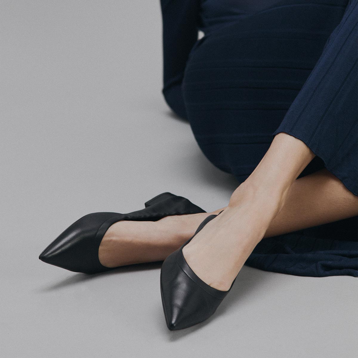 A person sits on the floor in a dark blue outfit, wearing Margaux's The Roma - Black Nappa flats with pointed toes, one leg crossed over the other against a plain gray background.