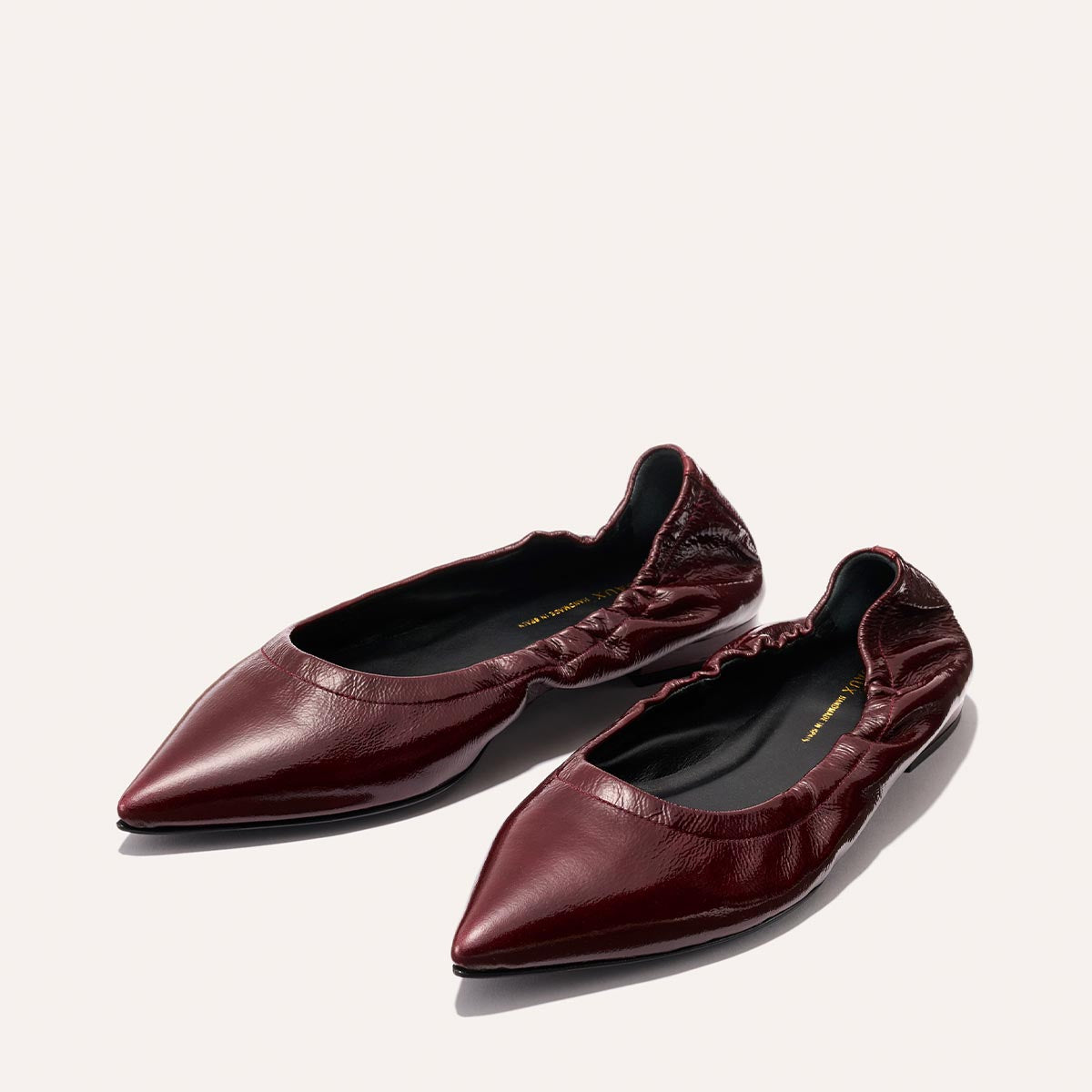 A pair of The Roma - Merlot Crinkle Patent ballet flats by Margaux, featuring a shiny burgundy finish and ruched elastic details around the opening, are displayed on a light background; these sophisticated shoes offer both style and elegance.