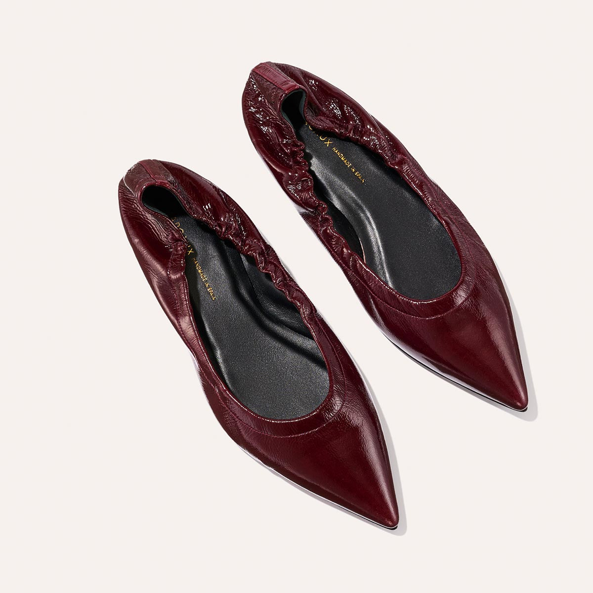A pair of Margaux’s The Roma - Merlot Crinkle Patent flats, featuring a burgundy pointed-toe design with elasticized backs, are displayed on a light background, blending sophistication with comfort.