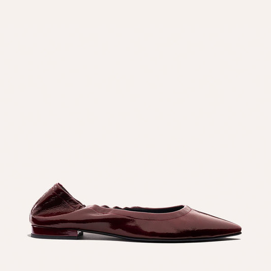 A side-view shot of The Roma - Merlot Crinkle Patent by Margaux, a sophisticated ballet flat in polished burgundy patent leather featuring a pointed toe, gathered elastic heel, and a low, slim black heel, set against a plain off-white background.