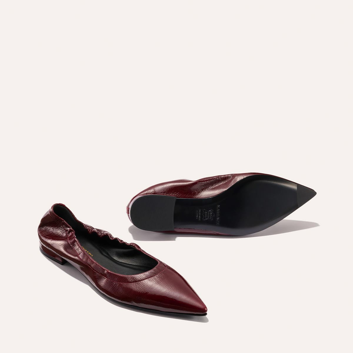 Two Merlot crinkle patent "The Roma" ballet flats by Margaux are displayed on a light background; one shoe is upright while the other reveals the sole, showcasing their sophisticated style and comfortable fit.