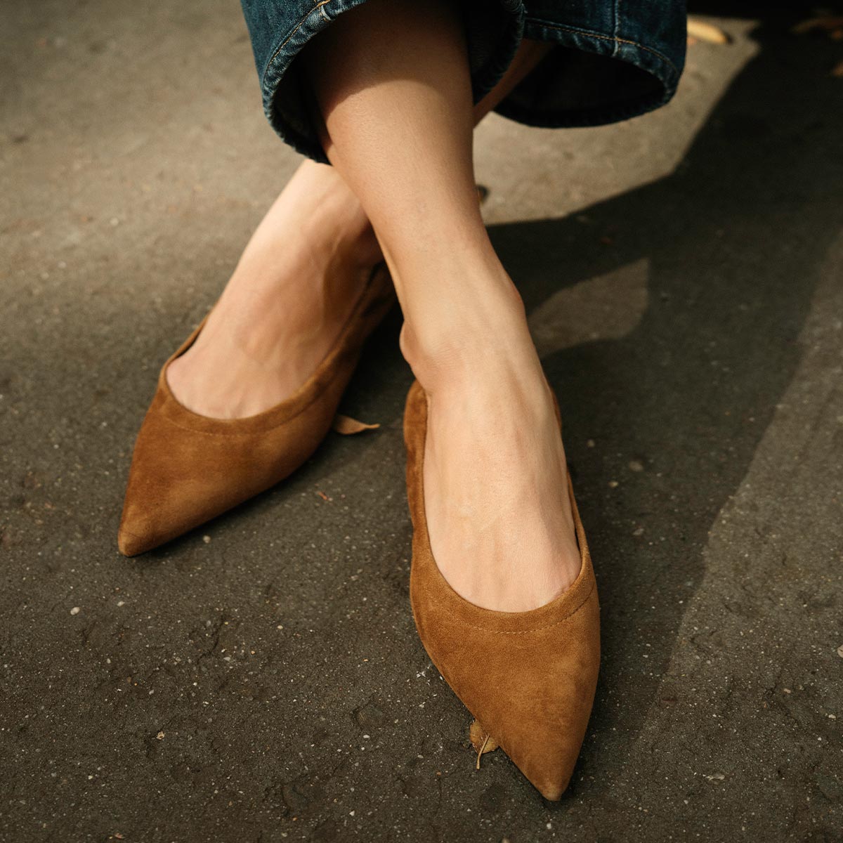 A person wearing Margaux's The Roma - Cedar Suede pointed-toe ballet flats with cropped blue jeans stands on a sunlit pavement.