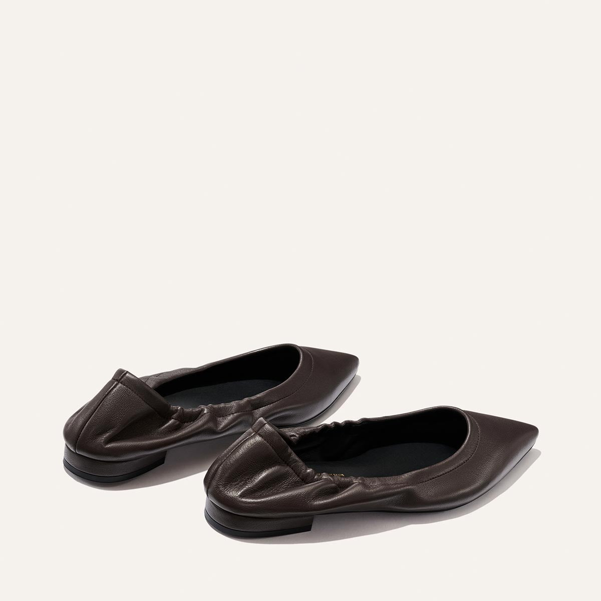 A pair of Margaux “The Roma - Espresso Nappa” sophisticated ballet flats in dark brown, featuring a pointed-toe design, ruched detailing, and low black heels, displayed on a plain light background.