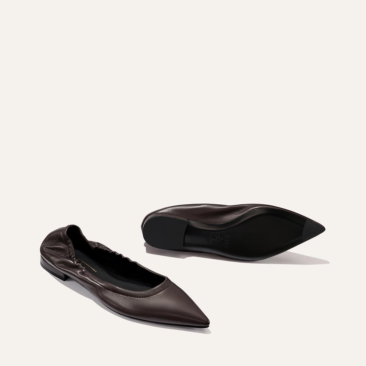 Two dark brown pointed-toe ballet flats from Margaux, called The Roma - Espresso Nappa, are shown—one upright and one lying on its side to display both the side and sole of the shoes. A light, neutral background highlights these sophisticated flats, making them perfect for any occasion.
