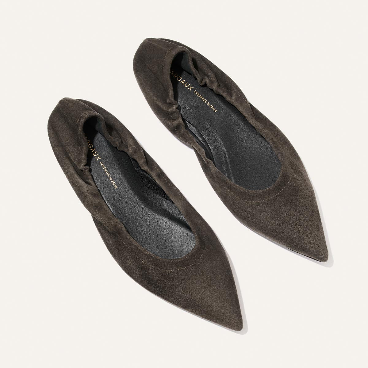 A pair of The Roma - Graphite Suede ballet flats from Margaux, featuring pointed toes and ruched edges in dark brown suede, are displayed against a light background.