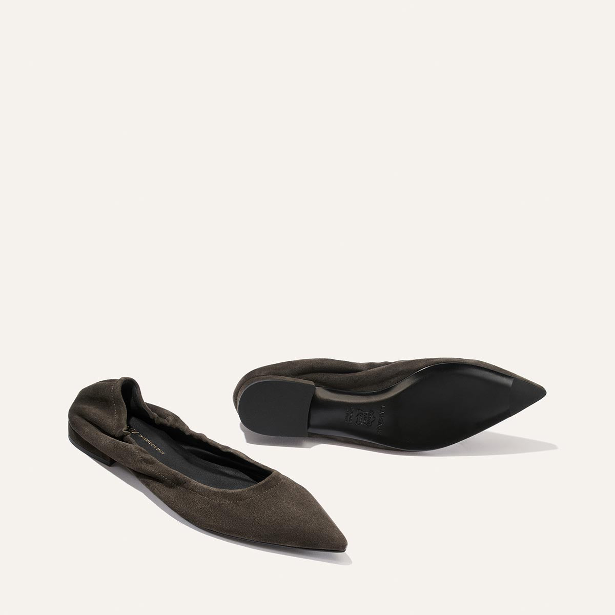 Two Graphite Suede “The Roma” ballet flats by Margaux—one positioned to reveal the elegant side and top views, the other turned over to showcase the black sole—rest on a simple light background, exemplifying an effortlessly sophisticated design crafted for both comfort and style.