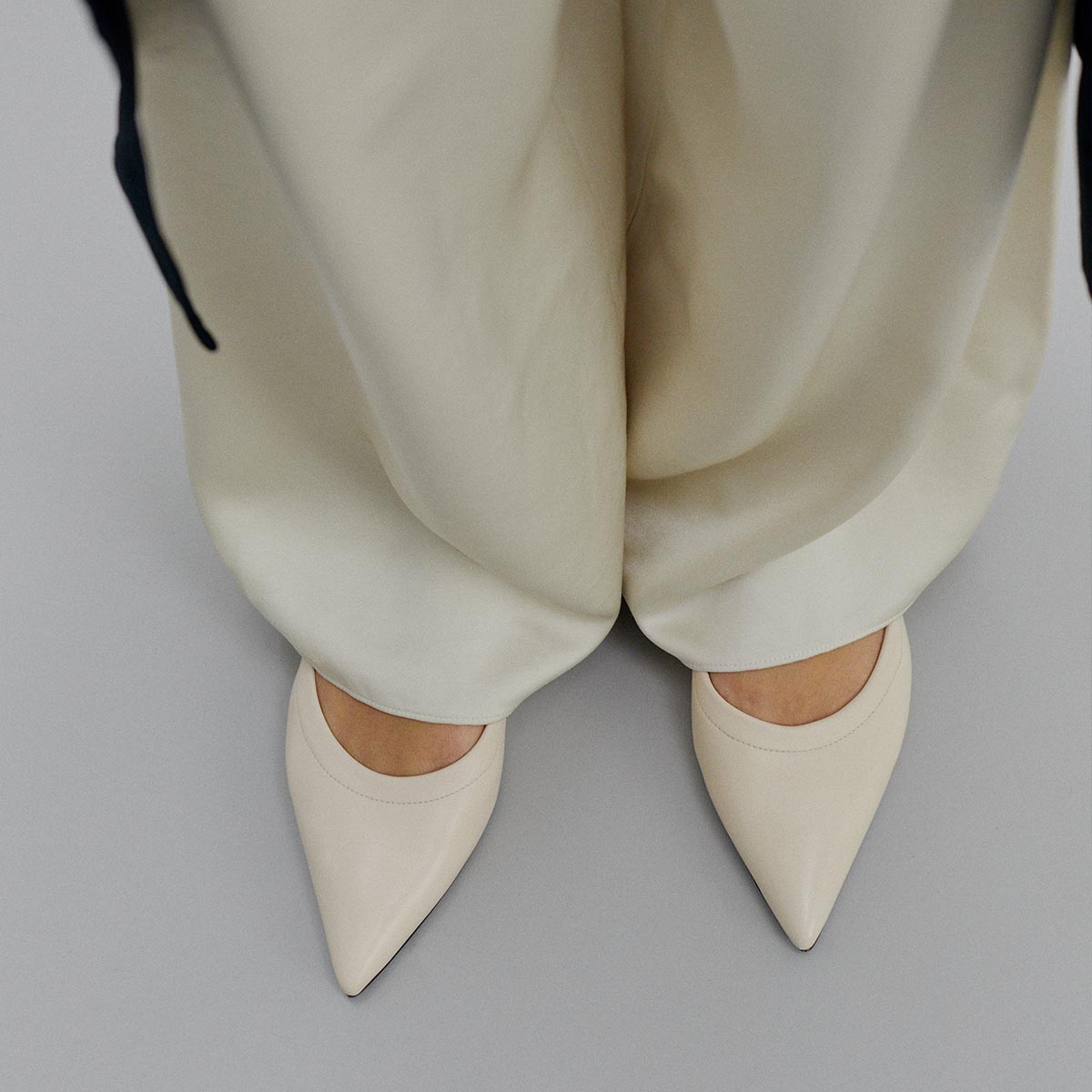 A person is standing on a light gray floor, wearing loose cream pants and Margaux’s The Roma - Ivoire Nappa ballet flats with pointed toes, viewed from above.
