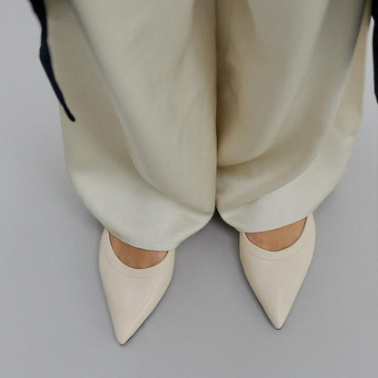 A person is standing on a light gray floor, wearing loose cream pants and Margaux’s The Roma - Ivoire Nappa ballet flats with pointed toes, viewed from above.