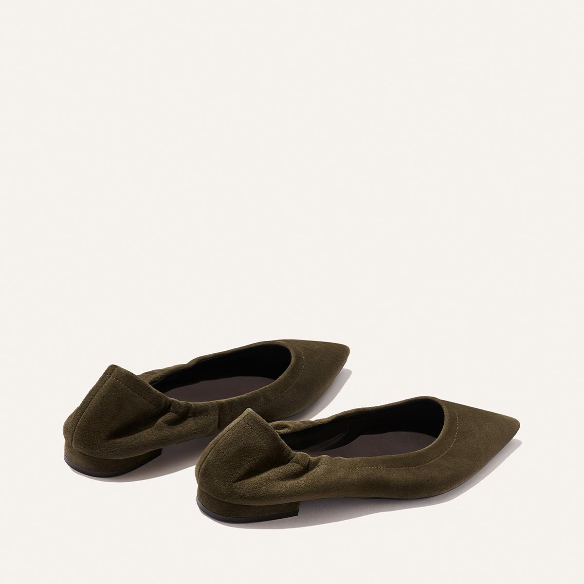 A pair of Margaux's The Roma flats in moss suede, featuring a pointed toe and ruched detailing, are displayed on a light beige background with the back and sides of the shoes visible.