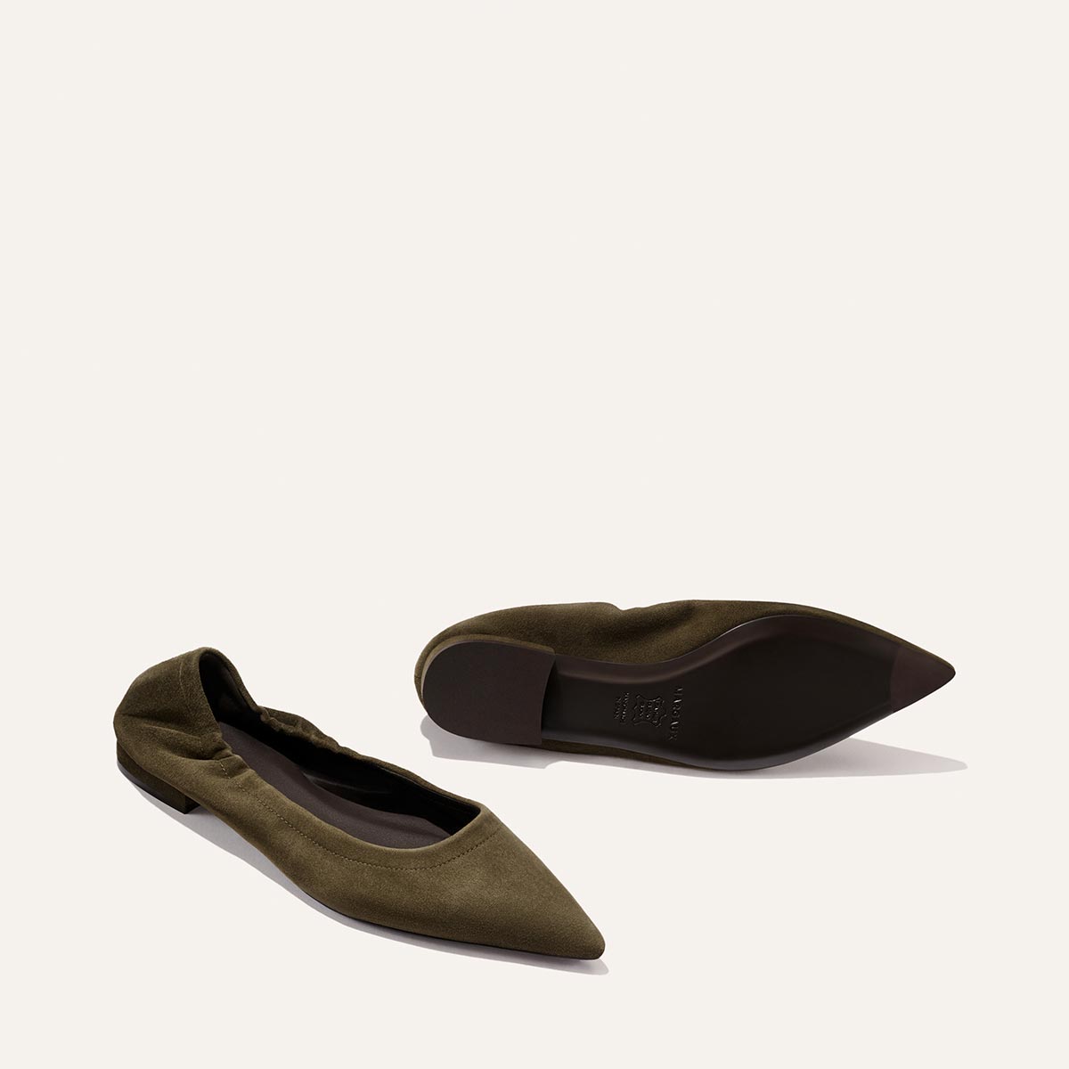 A pair of The Roma - Moss Suede flats by Margaux are shown against an off-white background: one olive green, pointed-toe shoe with ruched detailing is upright to reveal the top, while the other rests on its side to display the sole.
