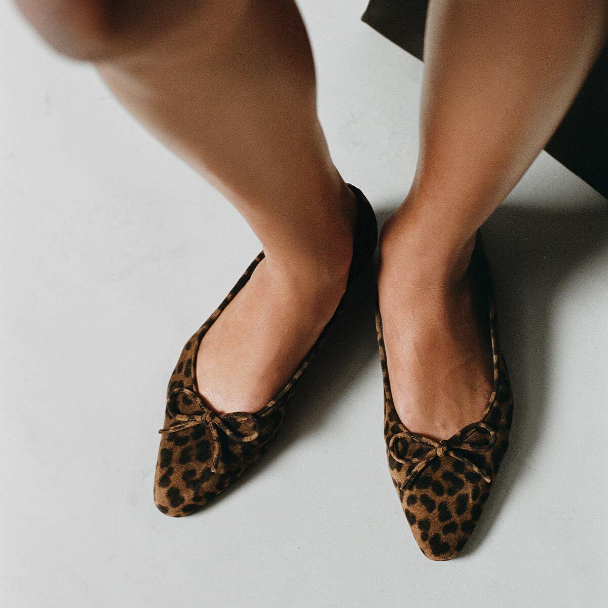 A person wearing The Salamanca - Brown Leopard Suede flats by Margaux, featuring pointed toes and small bows, stands on a light-colored floor.