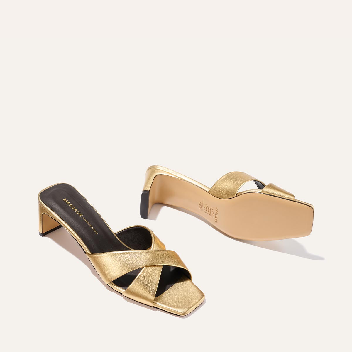 The Margaux Selene 50 - Gold Nappa sandals feature gold, open-toe criss-cross leather straps and a low block heel. Shown on a light background—one upright, the other on its side highlighting the sole.