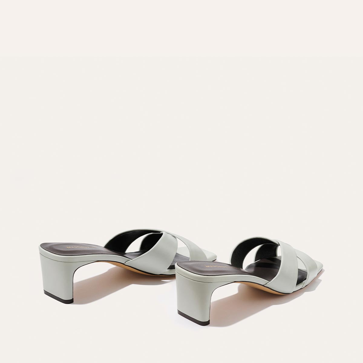 The Selene 50 - Sky Nappa by Margaux: light gray open-toe heeled sandals featuring crisscross leather straps, a square block heel, and a minimalist design, shown against a plain off-white background.