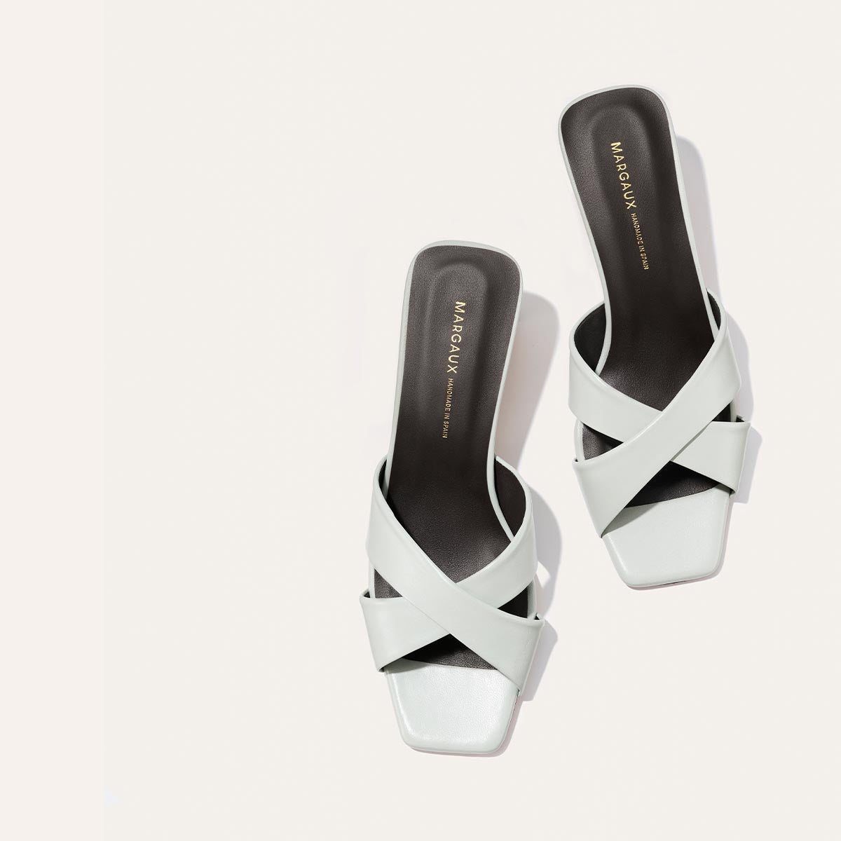 The Selene 50 - Sky Nappa by Margaux, featuring white open-toe slip-on heeled sandals with crisscross leather straps, square toes, and dark insoles with gold Margaux branding, are displayed side by side on a light background.