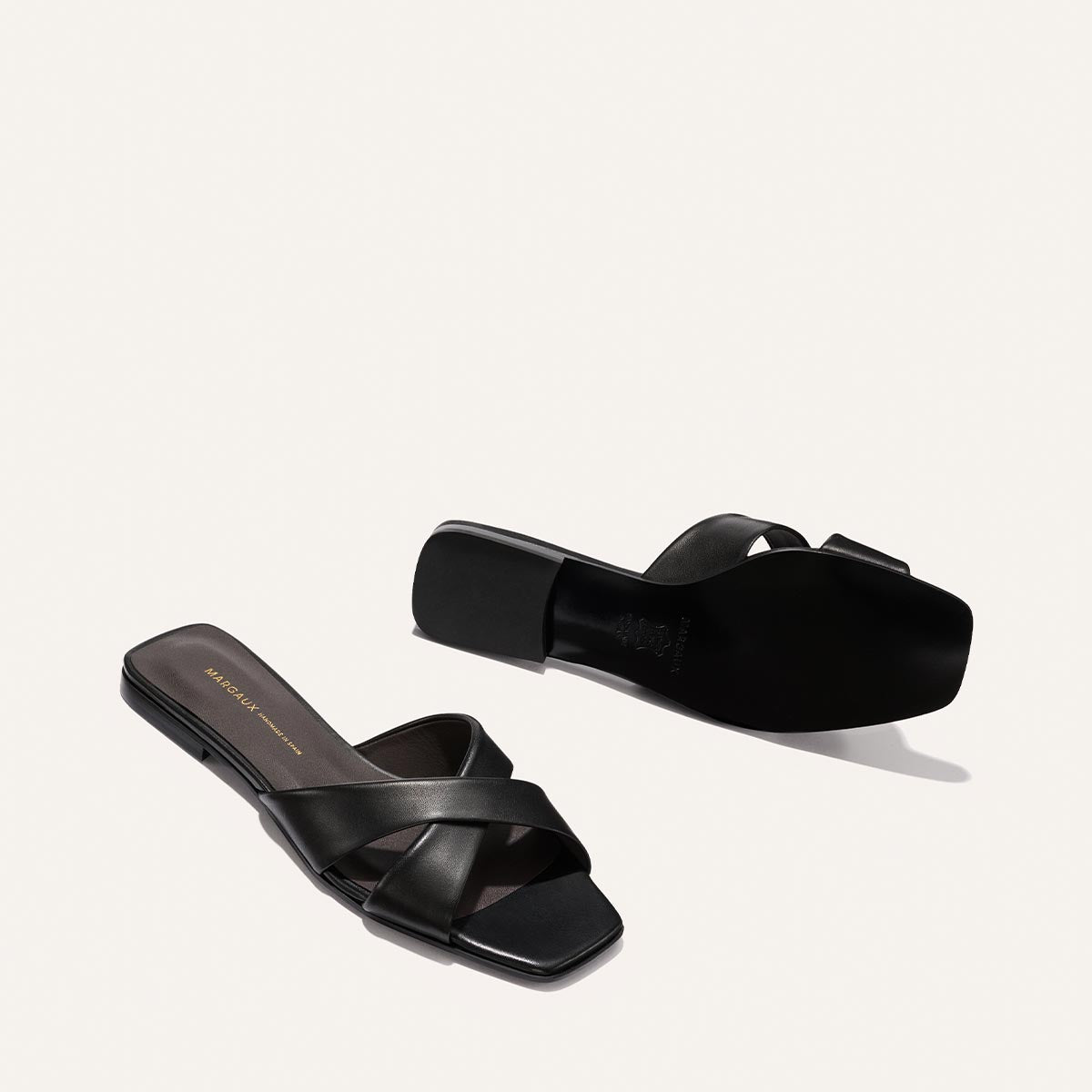 The Selene Flat Sandal - Black Nappa by Margaux is shown against a light background. One sandal stands upright, the other is flipped to display the sole. Features crisscross straps, square toes, and a slip-on design.