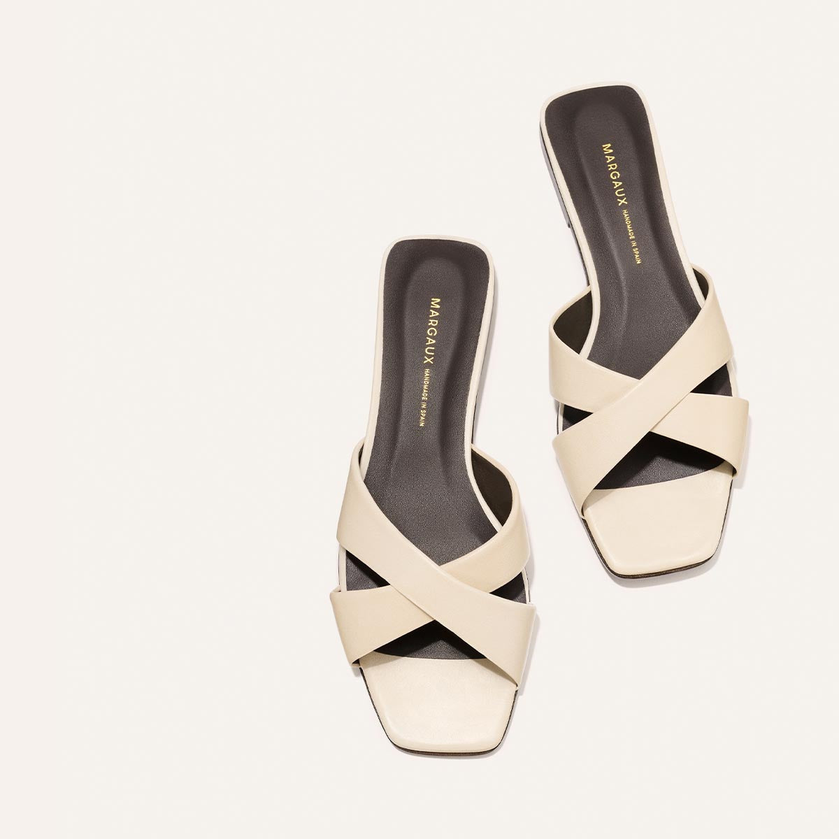 The Selene Flat Sandal - Ivoire Nappa by Margaux features beige open-toe crisscross straps, square toes, and dark insoles with gold Margaux branding.