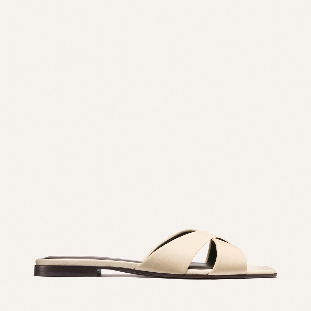 The Selene Flat Sandal - Ivoire Nappa by Margaux features a beige crisscross strap over the toes and a slim dark sole, shown against a plain light background.