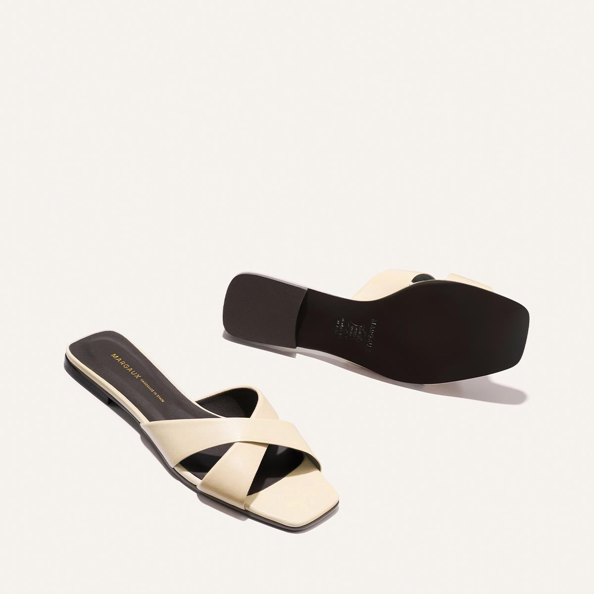 The Selene Flat Sandal - Ivoire Nappa by Margaux: a pair of beige open-toe sandals with crisscross straps and black soles, shown on a white background—one upright, the other flipped to display the sole.