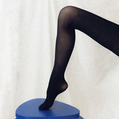 A single leg and foot in Margaux Semi-Sheer Tights - Black is shown resting on a blue, angular surface against a light, textured backdrop.