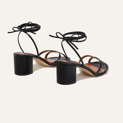 The Soho Sandal - Black Nappa by margaux-development is a pair of black open-toe block heel sandals with wraparound ankle straps tied in bows, displayed on a light background—an ideal summer staple.