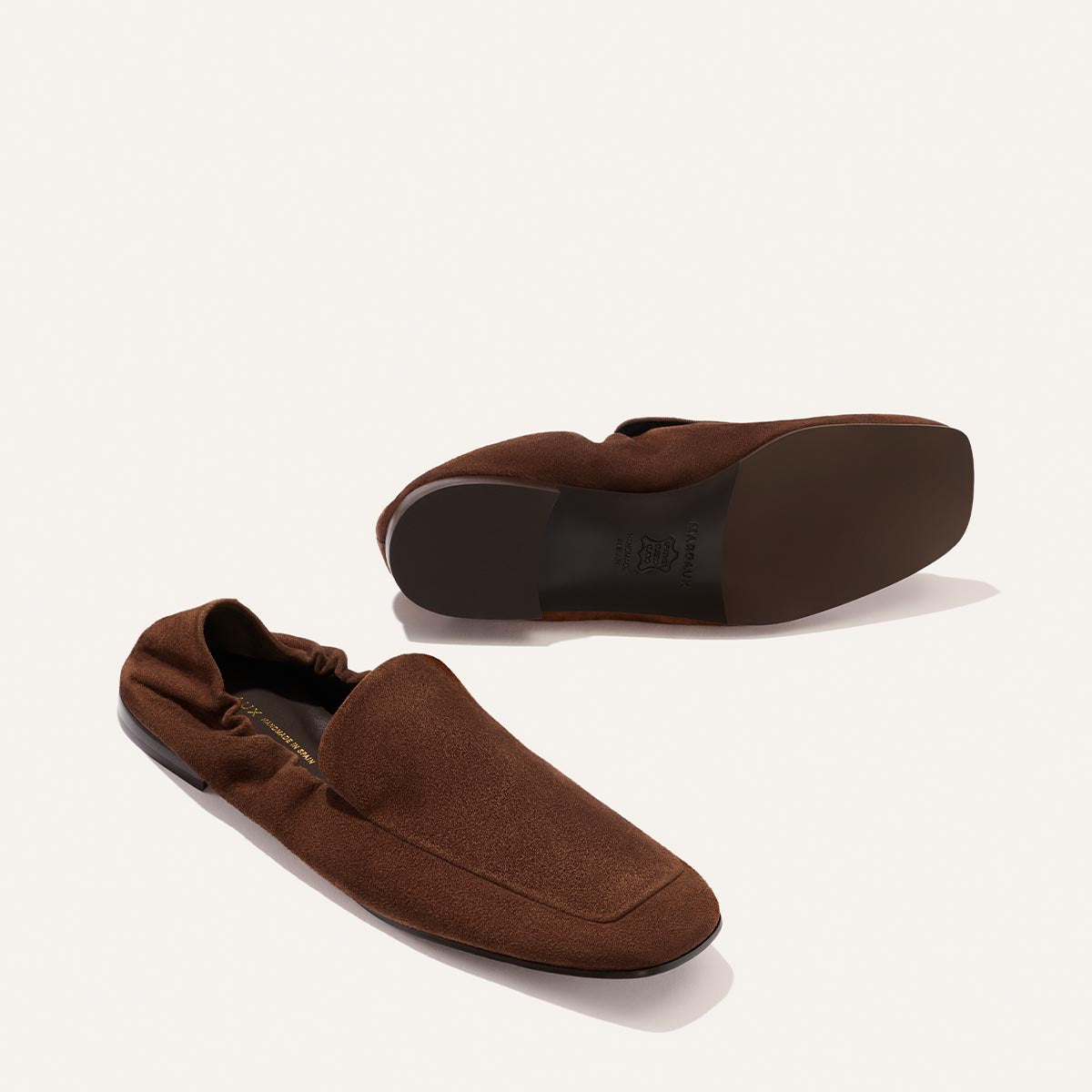A pair of Margaux's The Studio Loafer in Ganache Suede is displayed on a white background; one shoe stands upright while the other lies on its side, revealing the sole.