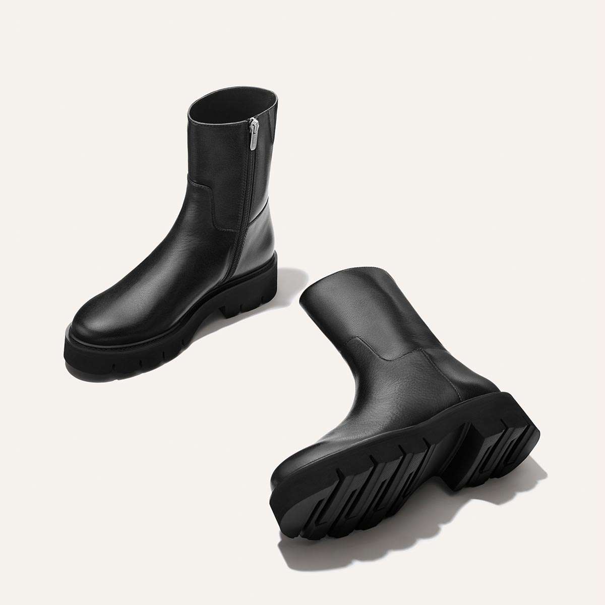 The Wells Boot - Black Calf by Margaux is featured against a light background, with one black leather ankle boot standing upright and the other lying on its side. Both boots have chunky rubber soles and side zippers.