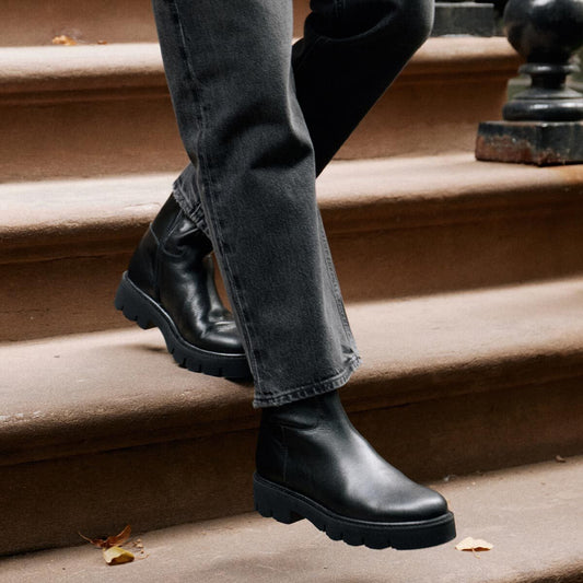 A person wearing black leather Margaux The Wells Boot - Black Calf ankle boots with lightweight lug soles while walking down stone steps on an all-weather travel day.