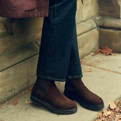 A person wearing dark jeans and margaux-development's The West Chelsea Boot in Cocoa Suede, featuring a sturdy lug sole, stands on a leaf-strewn sidewalk beside a stone wall.