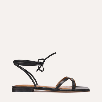 The Wrap Sandal - Black Nappa by Margaux features a sleek black wrap design with slender ankle ties, two minimalist toe straps, and a flat sole—ideal for versatile summer style. Shown in side profile against a plain background.