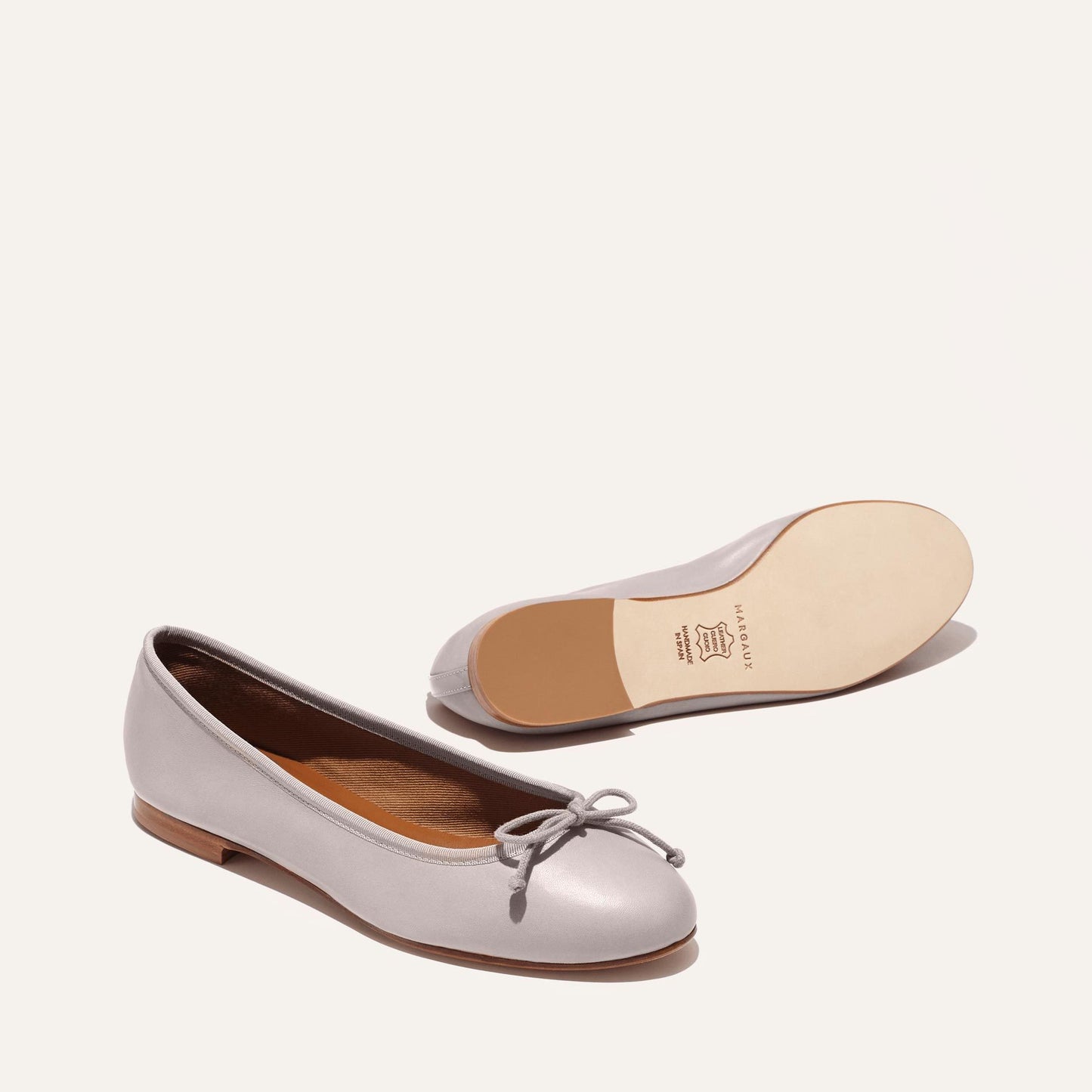 The Demi - Dove Nappa by margaux-development: light gray ballet flats, handmade in Spain with leather soles. One shoe displays the top and bow detail, while the other shows the sole and heel against a plain off-white background.