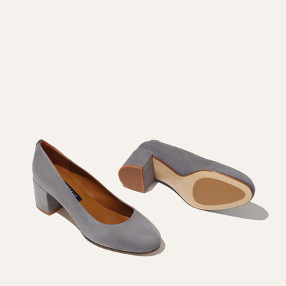 Margaux’s The Heel in Steel Suede—one elegant block-heeled pump stands upright to show the top and side, while the other lies sideways revealing sole and heel, both displayed against a plain off-white background.