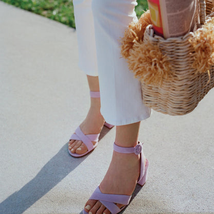 A person wearing margaux-development’s The City Sandal in Lilac Suede with a cushioned insole and white pants stands on a sidewalk, holding a woven straw bag with tan pom-poms. Grass shows in the background.