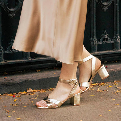 A person wearing a silky light gold skirt and The City Sandal - Champagne Nappa by margaux-development, featuring block heels, ankle straps, and cushioned insoles, stands on a sidewalk scattered with yellow leaves.