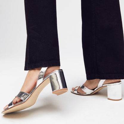 A person wearing margaux-developments The City Sandal - Silver Mirror, featuring a cushioned insole and two straps, paired with black pants, stands on a white background.