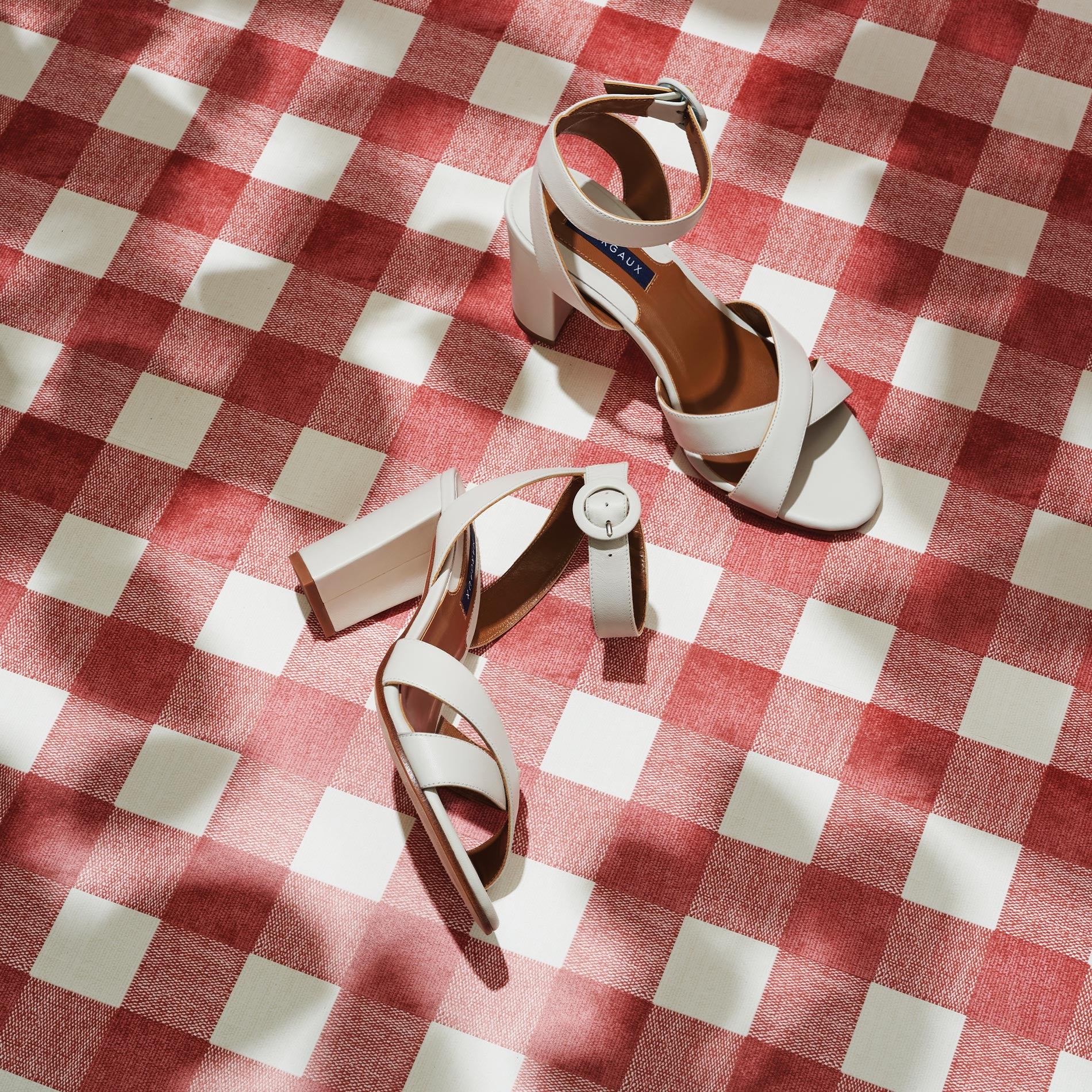 The Uptown Sandal - Ivory Nappa by margaux-development and a white wristwatch rest on a red and white checkered surface, sunlight creating soft shadows across the scene.