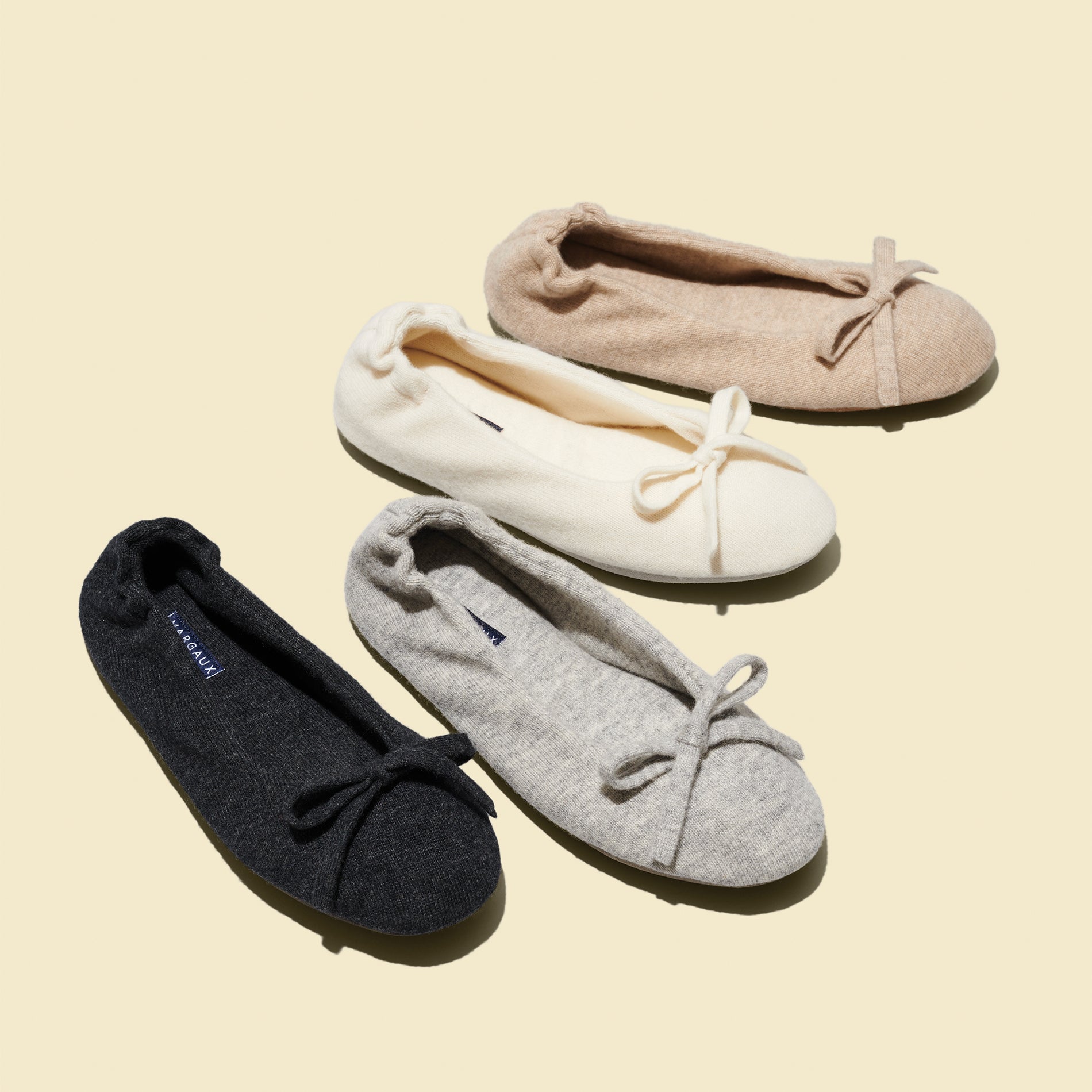 Four pairs of Margaux's The Ballet Slipper - Heather, styled like wool-cashmere ballerina flats with bows in black, gray, cream, and beige, are arranged diagonally on a light beige background. Perfect picks for the archive sale.
