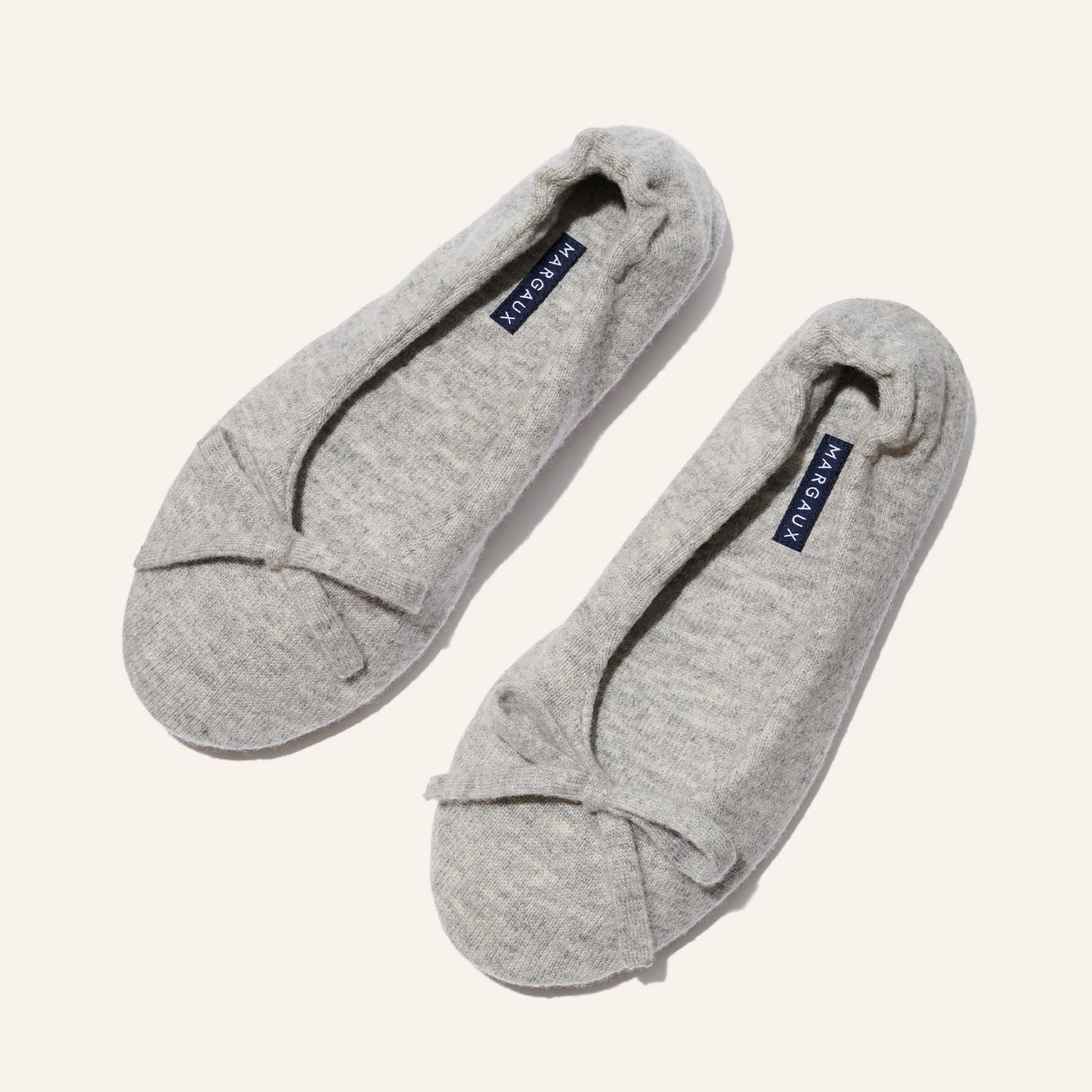 A pair of Margaux's The Ballet Slipper - Heather, cozy light gray slippers with soft fabric and small toe bows, is displayed on a neutral background. A dark blue label with white text decorates the insoles—an archive sale highlight.