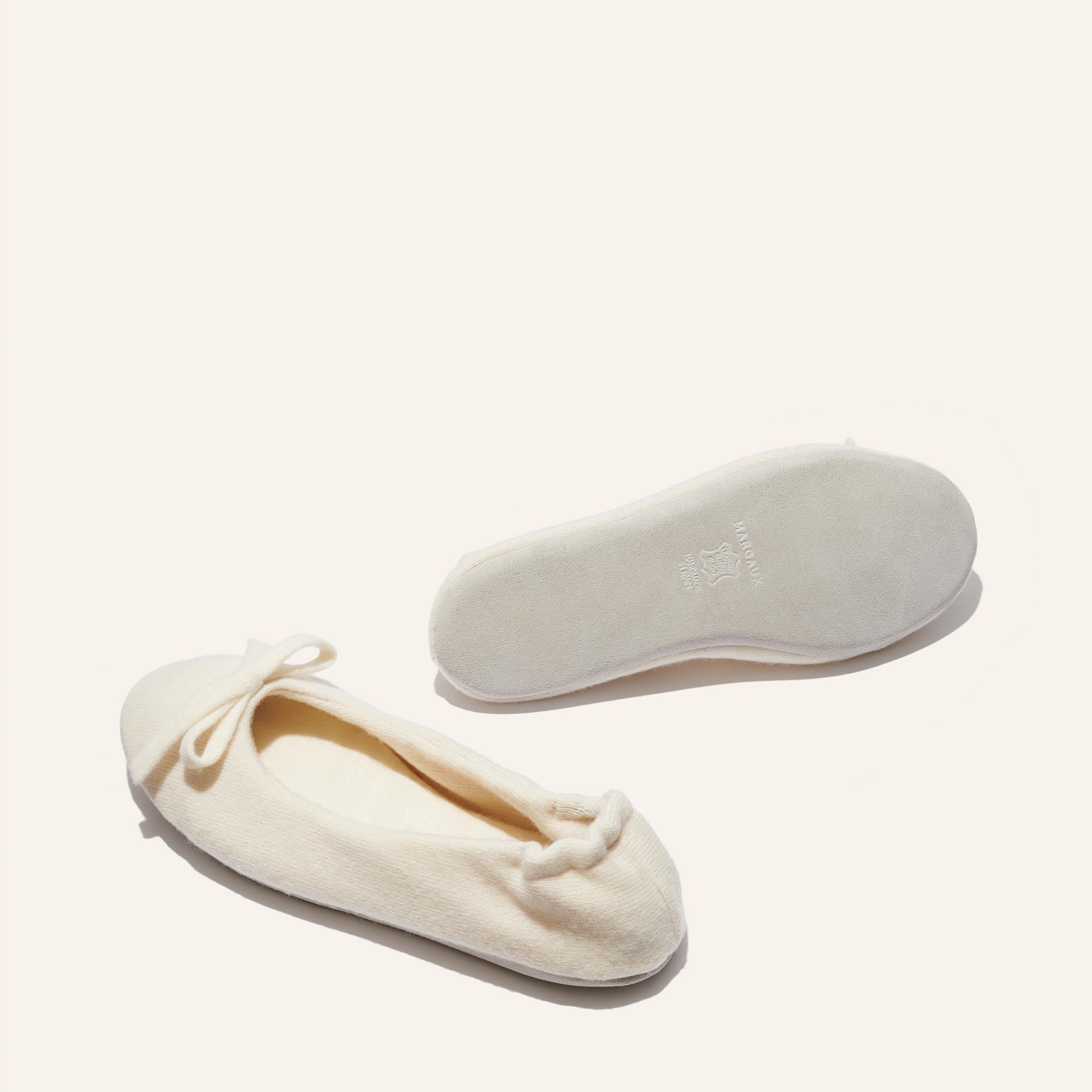 The Ballet Slipper - Ivory by Margaux: a cozy pair of cream ballet flats with toe bows. One shoe is shown from above, the other upside down revealing the sole, both set on a light beige background.