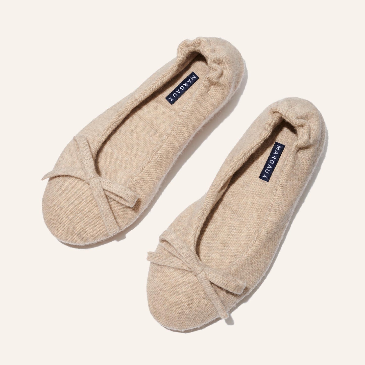 The Ballet Slipper - Oatmeal by Margaux features cozy beige ballet flats in soft, textured fabric with delicate toe bows and navy blue Margaux insoles—perfect for relaxing in style, elegantly presented on a light background.