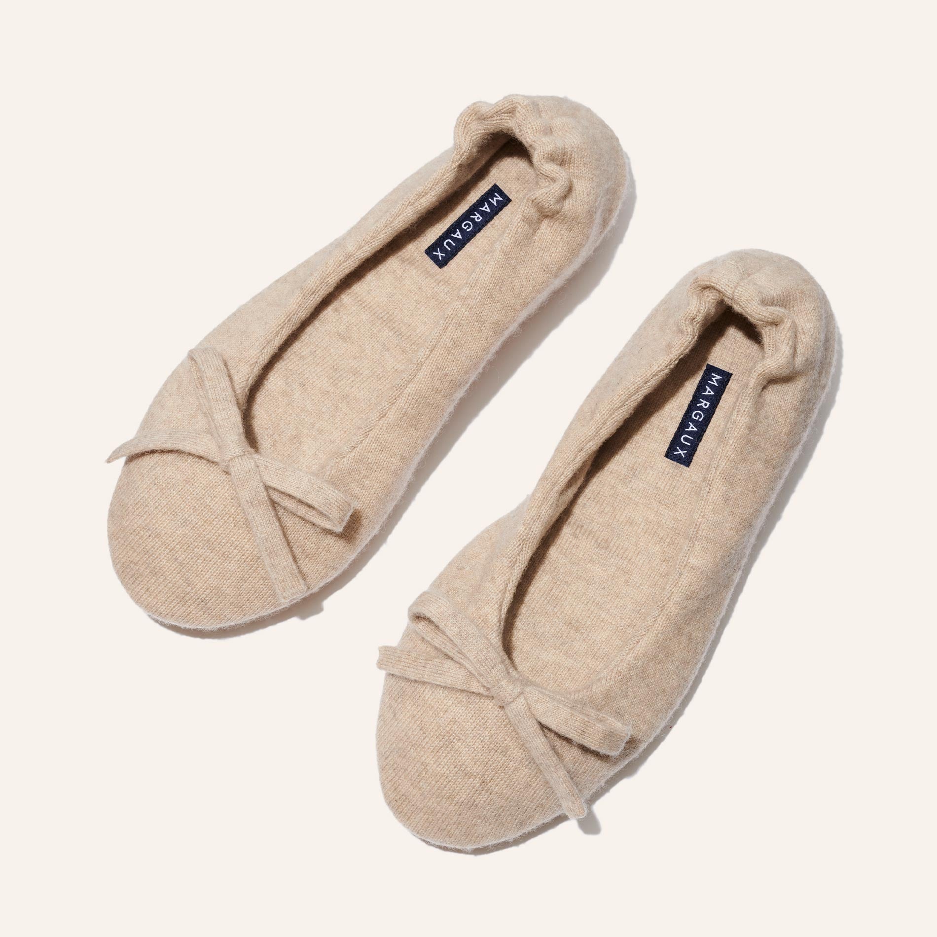 The Ballet Slipper - Oatmeal by Margaux features cozy beige ballet flats in soft, textured fabric with delicate toe bows and navy blue Margaux insoles—perfect for relaxing in style, elegantly presented on a light background.