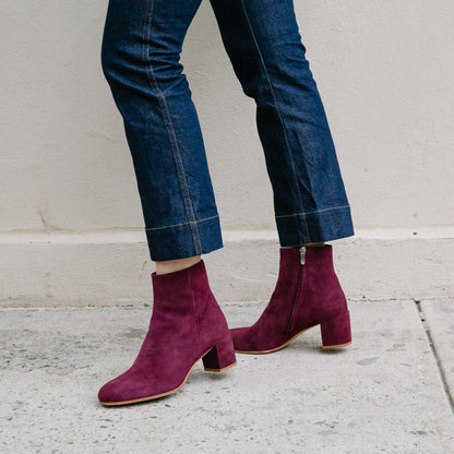 A person wearing dark blue cropped jeans stands on a sidewalk, showing off Margaux's The Boot in Mulberry Suede—stylish almond-toe boots with a block heel and side zipper.