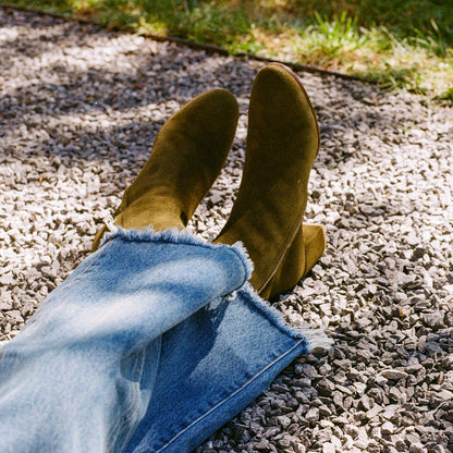 A person wearing frayed-hem blue jeans and Margaux’s The Boot - Olive Suede relaxes with crossed legs on gravel, with grass in the background.