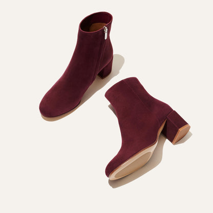 The Boot - Mulberry Suede by Margaux: One chic burgundy suede boot stands upright beside another on its side, revealing the sole. Both feature almond toes, block heels, and side zippers, set against a light background.